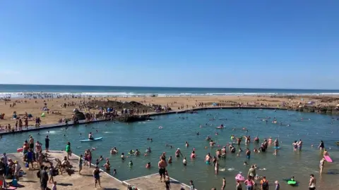 Bude Sea pool on a sunny, blue sky bank holiday Monday, with lots of people in the water swimming and paddling. The beach behind it separates the pool from the sea.