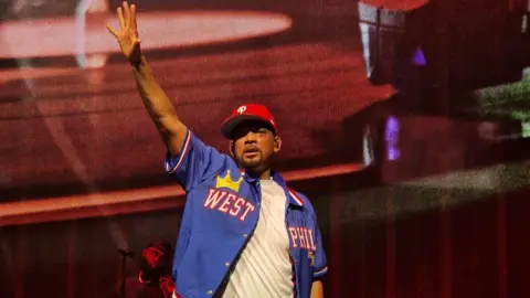 Kevin Tasker Will Smith in a white T-shirt and open blue baseball shirt with West Phil on it, and wearing a red baseball cap slightly twisted to the right. He is on stage and has his right hand raised in the air. He looks a little out of breath and sweaty. The stage behind him is backlit in red.