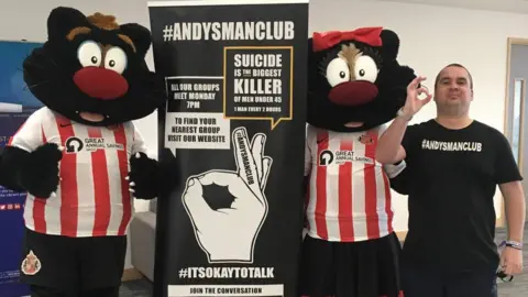 Supplied Michael Chapplow is wearing a black t-shirt reading ANDYSMANCLUB. He is standing next to banners about encouraging men to talk about their mental health. Two Sunderland AFC black cat mascots in the club's red and white t-shirts are standing on each side of the banner.