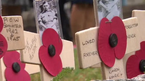BBC Wooden handmade crosses with pictures of men from the war, in the middle are paper poppies, all are written on with personal messages. They are sticking out of the ground.