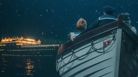 The backs of three people are seen in a lifeboat looking towards a large cruiseliner. It's night time and the lights are on in The Titanic.