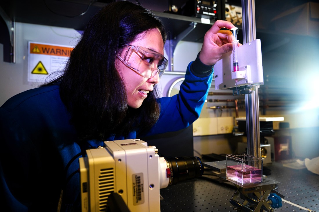 Xiaoyu wearing goggles and a blue lab suit examining a syringe of red liquid in the lab.