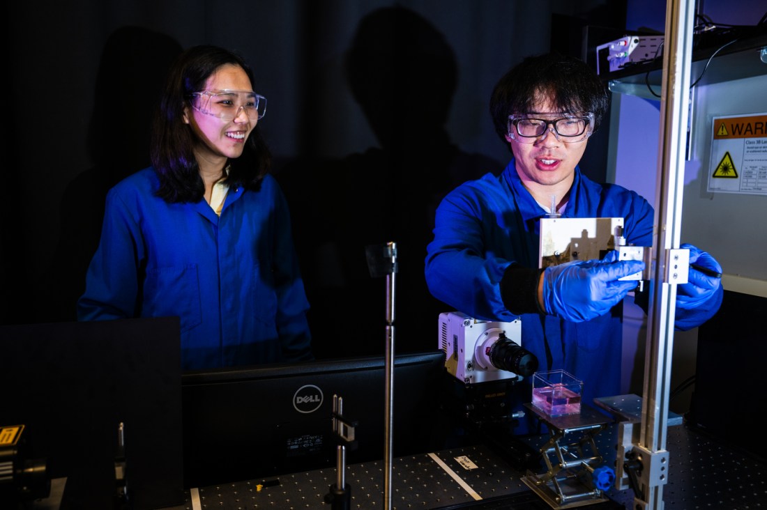 Xiaoyu Tang guides Boqian Yan in the lab in adjusting equpiment. Both are wearing goggles and blue lab suits.