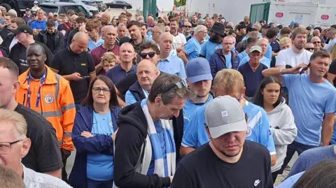 @Ticketmancity Large crowd of about a 100 people waiting. Most are wearing blue tops, including the City shirt. Many are looking frustrated or unimpressed, with some on their phones. A club steward in an orange stands to the left.