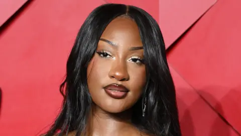 WireImage/Getty Images Tems looks past the camera, she has long, straight, black hair and stands in front of a red background