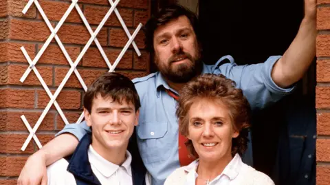 Lime Pictures/Rex Features Bobby, portrayed by Ricky Tomlinson, and Sheila Grant, played by Sue Johnston are standing in front of their red-bricked home on Brookside Close with one of their children. They are all smiling at the camera in this posed photograph.