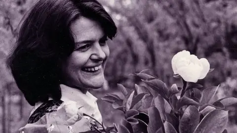 Maggie's Centres A black and white photograph of Maggie Keswick Jencks. She has shoulder-length, dark hair. She is only visible from the shoulders up. She is carrying flowers in her arms, the top of which, with leaves and a single light-coloured bud, is visible.