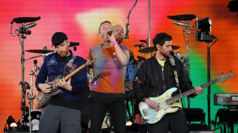 PA Media A band performs on stage with three members in the foreground: a guitarist on the left, a singer in the centre, and a bassist on the right. Colourful stage lighting in red, orange, and green fills the background, with drums and equipment visible behind them.