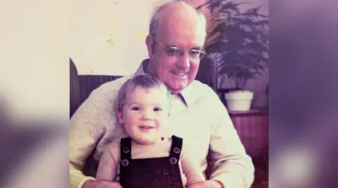 An old photograph of an elderly man with a young boy on his lap. The elderly man is balding and has a pair of reading glasses on. He is wearing a light coloured jumper and a collared shirt under it. The little boy has short hair and is wearing dark dungarees. Both of them are looking at the camera and smiling. 