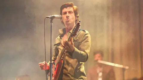 Getty Images A man with brown wavy hair on stage, standing behind a microphone and holding a guitar. He is looking into the crowd.