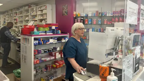 BBC A pharmacy with a member of staff wearing a blue uniform at a till standing in front of a cabinet full of medicines.