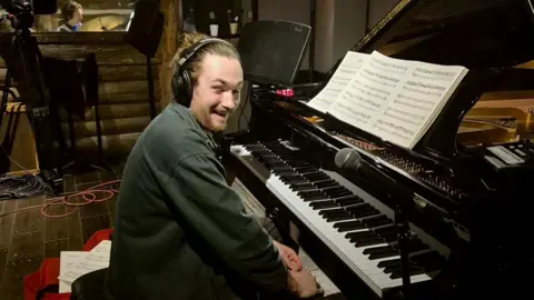 Fergus McCreadie A man laughing as he sits at the piano in a recording studio, with sheet music in front of him