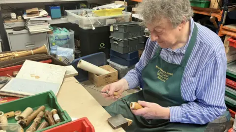 Daniel Bangham inserts a long thin metal tool into a wooden cylinder held in his other hand. Daniel has grey hair and is sitting at a work table, wearing a blue and white striped long-sleeved shirt and a dark green apron with yellow writing on it which says: Woodwind & Reed Cambridge Woodwind makers. Woodwind pieces, similar to what he is holding, are in a green plastic tray on the table.