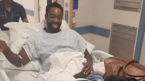 Handout Matthew Allick in a hospital robe and on a stretcher inside a hospital. He smiles at the camera.