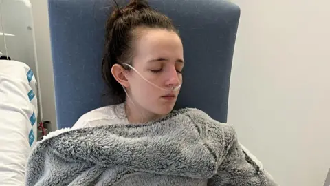 Amy Symington Amy Symington is sitting in a high-backed chair in hospital. She has an oxygen tube attached to her nose. She has a thick, grey fleece blanket wrapped around her and is dozing. To the left we can see the side of a hospital bed. 