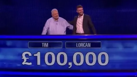 ITV Tim McCarthy and Lorcan Thorpe appear on The Chase. They are standing in the studio on the blue set behind a screen that shows their names and the prize money of £100,000.
