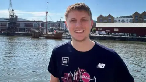 Family handout A head and shoulders shot of a man smiling at the camera wearing a navy T-shirt with a design on. He has short brown hair. Behind him is a river, boats and buildings in the distance. The sky is blue.