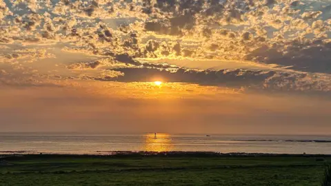 Bryan Wark Landscape image of the sun setting over calm waters at Turnberry beach. The sky is an orange, yellow and blue ombre with the sun peaking from behind clouds.