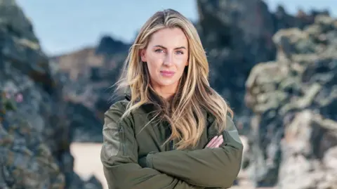Pete Dadds/Channel4 Michaella McCollum has long blonde hair and is looking at the camera with a neutral expression on her face as she folds her arms. She is standing on a beach with large, jagged rocks behind her. She is wearing a green zip-up jacket.