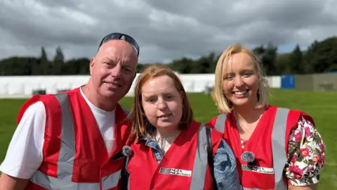 Three people are standing in pink high visibility vests. The man on the left is wearing a white t-shirt, he has black sunglasses on his head. The girl in the middle has a denim jacket on, she has red hair. The woman on the left is wearing a flowery top and has blonde hair. The background is blurry. 
