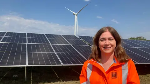 Amy Holmes/BBC A picture of Labour MP Alex Mayer, a woman with shoulder length dark brown hair in a centre parting. She is wearing a high visibility jacket and is stood in front of some solar panels.