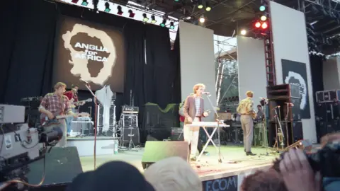 Mark Hodgson An outdoor stage with a large logo at the back of the stage reading 'Anglia for Africa', over a shape of Africa inside an outline shape of Norfolk and Suffolk. Performing on the stage is a pop group comprised of four young men, two playing guitars, one standing at a microphone while also playing a keyboard, and another man standing turned away from the camera. A few members of the audience are partly visible at the bottom right and bottom centre of the image, and a television camera pointing at the stage is visible bottom left.