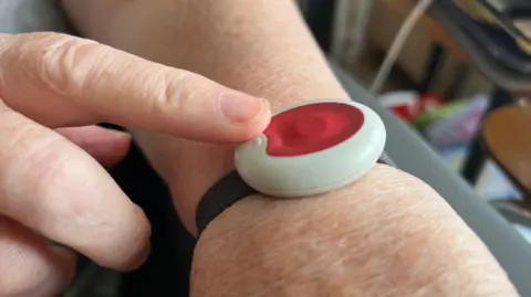 BBC Photo of a red alarm button strapped to a wrist with a black strap with a finger touching it