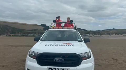 BBC Two young male lifeguards in red jackets stand on the back of a white RNLI branded truck. They are on a sandy flat beach, with hills behind.