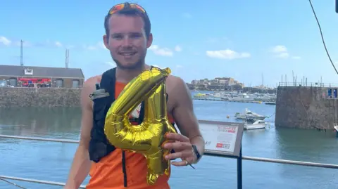 James Hadley-Piggin James stands near a waterfront holding a large gold balloon shaped like the number '4'. They wear an orange sleeveless top, black shorts, and a hydration pack. Behind them, boats are docked and buildings line the shore under a clear blue sky