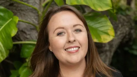 Kimberlyn Murphy A woman smiling straight at the camera. She is standing in front of a tree and palm leaves.