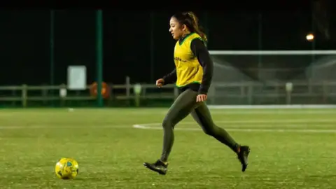 Simran Sandhu A women stands on a football pitch kicking a football. It is very dark and she is wearing a yellow T-shirt and black leggings