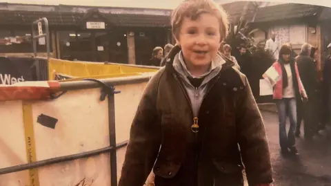 Handout Harry Churchill as a young boy at a farm event. He is wearing a dark zip up top with around three layers beneath of grey, navy and white in colour. He is smiling at the camera and is around aged 6. 