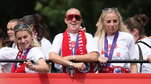 Reuters Three england footballers, wearing white tshirts and medals on purple lanyards around their neck, are looking over a black metal railing. 