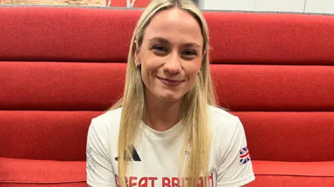 Izzy with long blonde hair sitting on an orange sofa smiling at the camera. She is wearing a white 'Team GB' T-shirt.