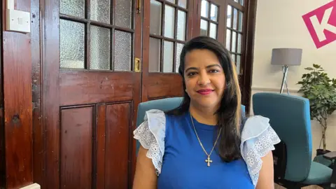 Ollie Conopo/BBC Melisha Pillay looks directly at the camera as she is photographed sitting in a room with a dark wooden door behind her. She is wearing a blue top with stripey, ruffled sleeves. She has a long gold chain around her neck with a cross hanging from it. She has dark hair. 