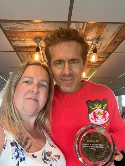 Sarah Waller Sarah Waller pictured with Ryan Reynolds, who holds a circle glass plaque with a piece of turf inside and with writing inscribed on top. Reynolds wears a red knit jumper with the Wrexham shield, has brown hair and brown eyes and smiles at the camera. Sarah has blonde mid length hair and wears a white and blue floral vest top and looks at the camera. 