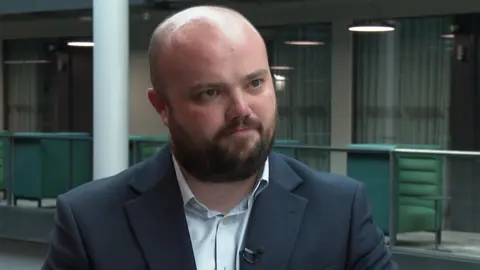 BBC Close up image of Llŷr Powell standing in the BBC building in Cardiff. He is wearing a light blue shirt and navy blazer which has a small microphone attached on the left collar. He is looking at the interviewer not the camera. Powell is bald with a brown beard and has a stern expression. 