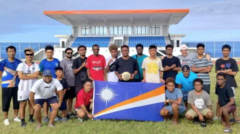 Marshall Islands Soccer Federation A team of young men pose on a pitch with a football and the flag of the Marsahall Islands - which is blue, with a white star and diagonal white and orange stripes. There is a small sports stand behind them and the sea beyond.