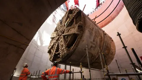 HS2 A huge section of a digging machine is being winched out of a tunnel watched by two workers in orange high vis clothing and hard hats.