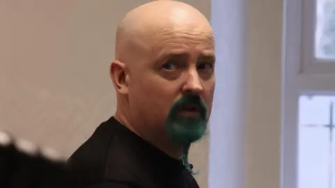 Emily Frances Phil Churchill. He is bald and has a beard that's dyed turquoise. He is frowning slightly and looking to the side.