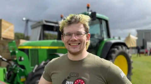A man is wearing a green t-shirt. He has black and blonde hair. He is also wearing glasses. In the background there is a green tractor. 