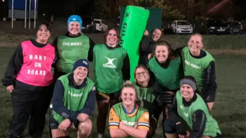 Trish Brown A group of women rugby players all in green vests except Trish, whose is pink and says 'Baby on Board' on it