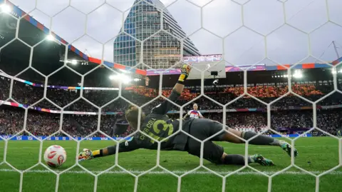 PA Media a photo taken through a goal net. In the foreground, a keeper in dark green kit dives to her left, unable to make contact with a football. In the background, the top half of a footballer in an England shirt is visible. Behind her are packed stands on all sides, and a large skyscraper in the background.