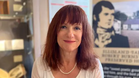 Joan McAlpine, a red-headed woman wearing a white top and pearls, smiles sitting in the museum with a Burns information board behind her.