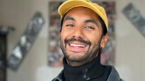 Jay Singh wearing a black knitted turtleneck jumper, a thin gold hoop earring and a bright yellow cap. He is smiling at the camera and the background is blurry.