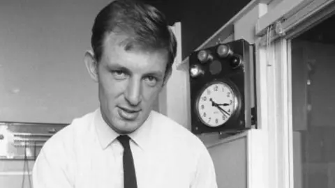 A black and white photo of Gerry Harrison, dressed in a white shirt and thin black tie, with a window to the side of him and a clock with light bulbs behind him.