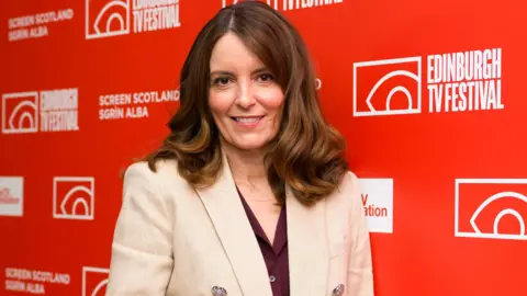 PA Media Tina Fey, who has log brown hair and is wearing a maroon blouse with a beige blazer, standing against a bright red backdrop multiple white logos including "Edinburgh TV Festival" and "Screen Scotland"