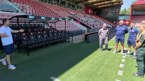 Tom Lockyer is wearing a white T-shirt with blue shorts and white running shoes with pink and orange-trimmed soles. He is walking out on to a football pitch of an empty stadium with his arms outstretched towards two paramedics and three men in football club training gear. They are standing in the sun on the edge of the manager's technical area - signified by a white dotted line painted on the grass - while Tom is in the shade cast by the stand above him
