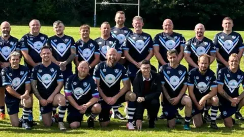 Redruth Albany RFC A team photo of the rugby club's players lined up in two rows on the pitch with rugby posts behind them. The first row is kneeling and the second row standing and the players are all clasping their hands together in front of them.