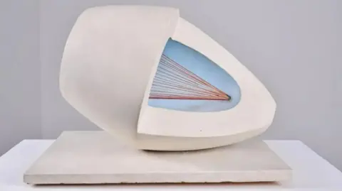 Betty Saunders A white oval sculpture with pale blue and red colours in the middle. It has been placed on a white slab for display.